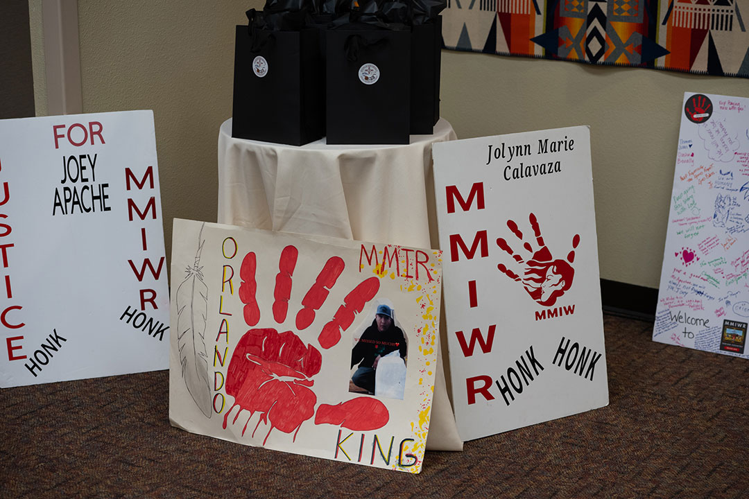 _DSC4727 Hand-painted posters honoring missing and murdered Indigenous relatives, including signs reading Justice for Joey Apache, Orlando King, and Jolynn Marie Calavaza, each featuring a red handprint, displayed in front of a table with black gift bags.