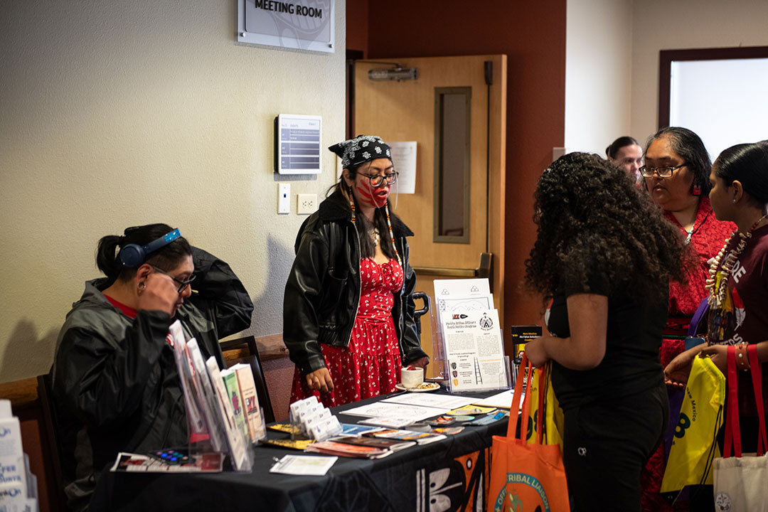 DSC_6101 Attendees gather around an information table at an MMIWR awareness event, where a woman wearing a red dress and a red handprint painted across her face speaks with visitors holding tote bags.