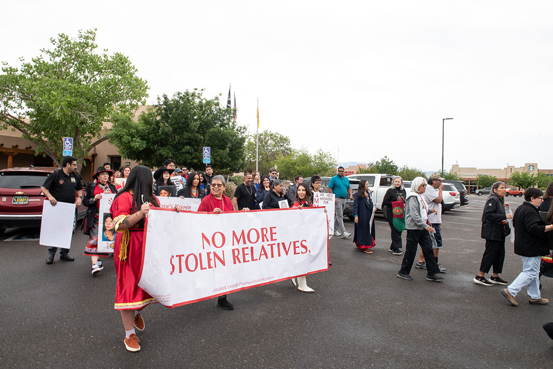 DSC_6197 Marchers walk through a parking lot during an MMIWR awareness walk, led by participants in red carrying a banner that reads No More Stolen Relatives.